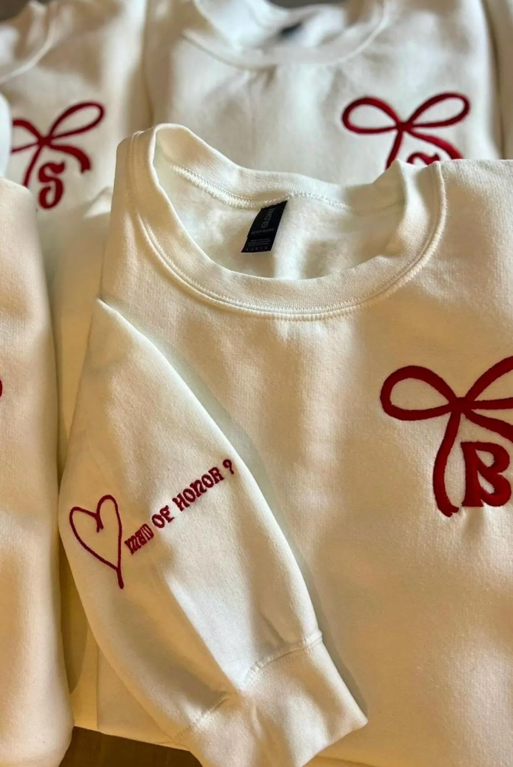 A collection of crewneck sweatshirts with embroidered bows and initials on them, displayed in a pile.