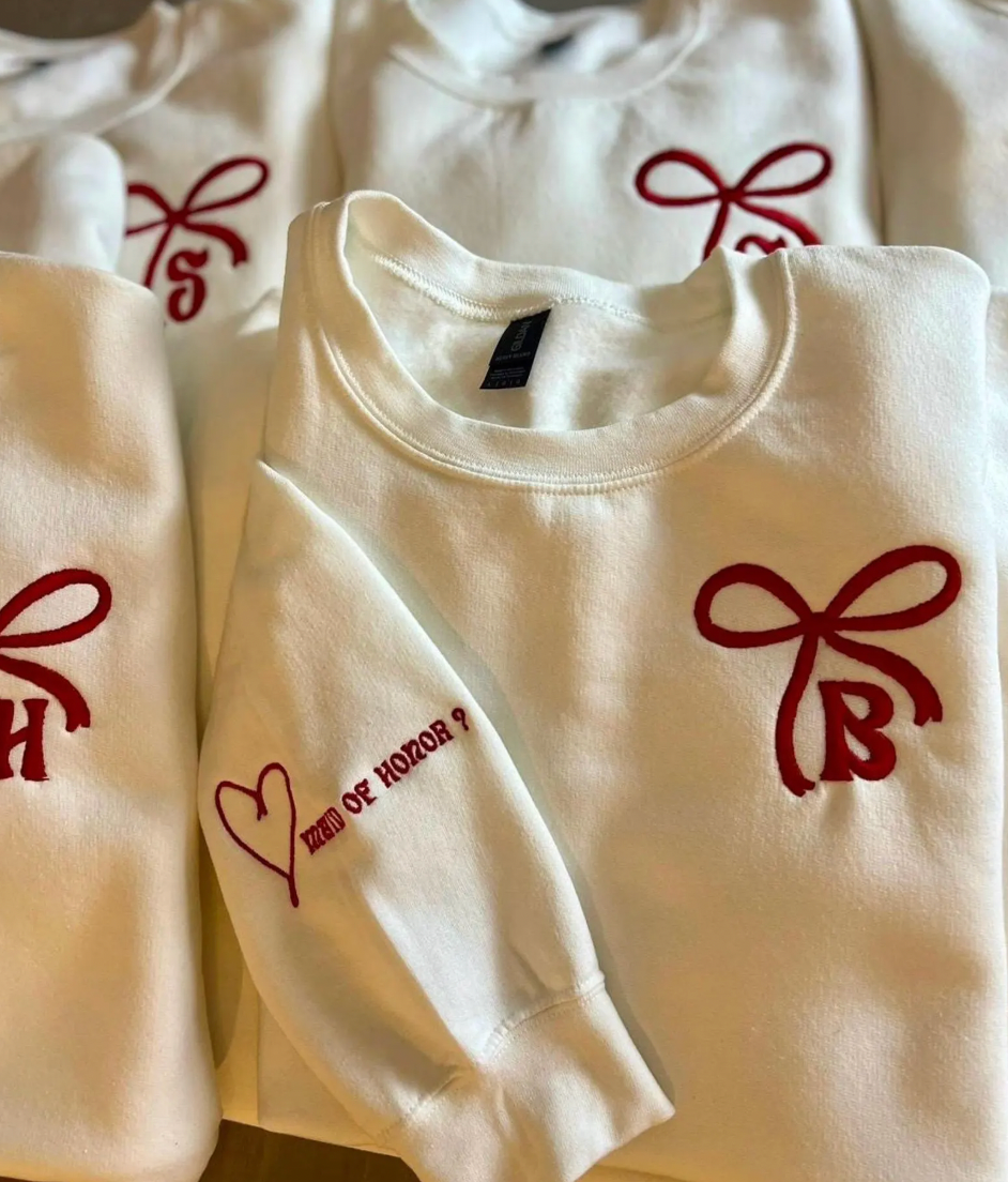 A collection of crewneck sweatshirts with embroidered bows and initials on them, displayed in a pile.