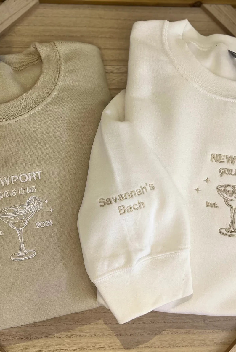 Two folded embroidered crewneck sweatshirts, one beige and one white, with a custom margarita glass design and text on them.