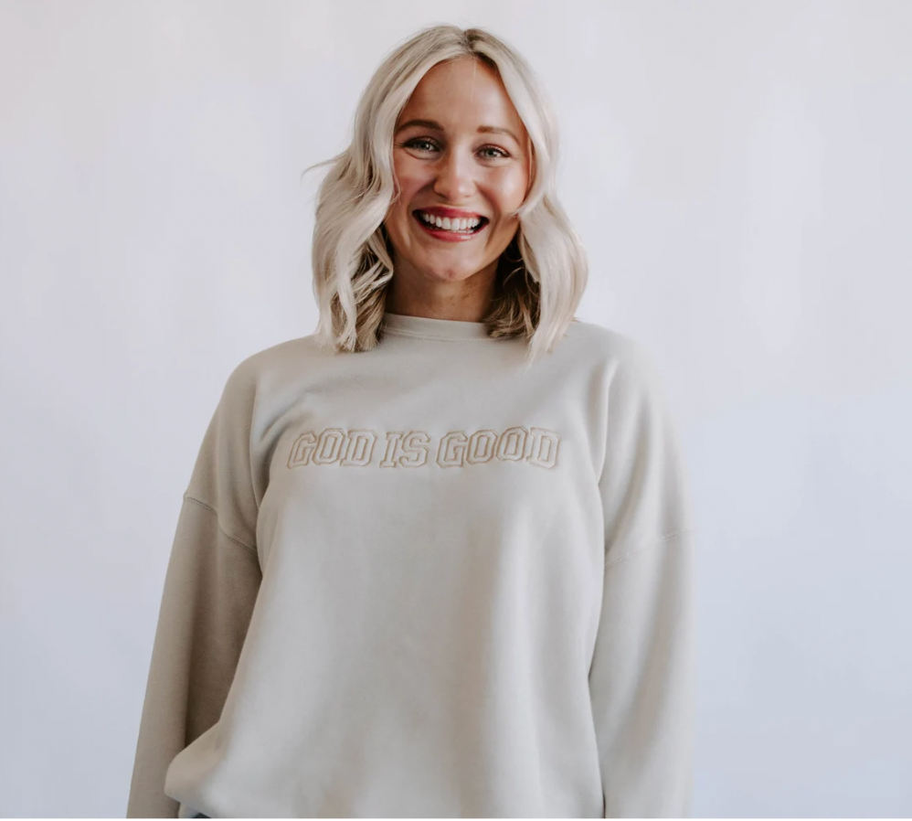 A person wearing a dust-colored sweatshirt with 'GOOD IS GOOD' embroidered on the front.