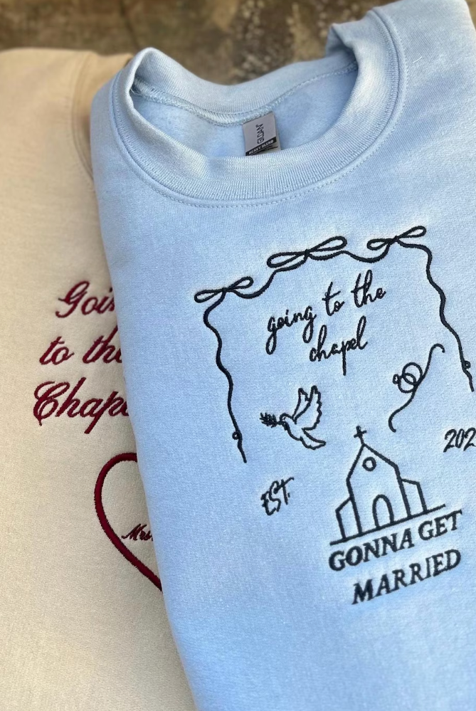 Light blue embroidered sweatshirt with text and design elements indicating a bridal theme, displayed next to a beige item, possibly another sweatshirt or a hat.
