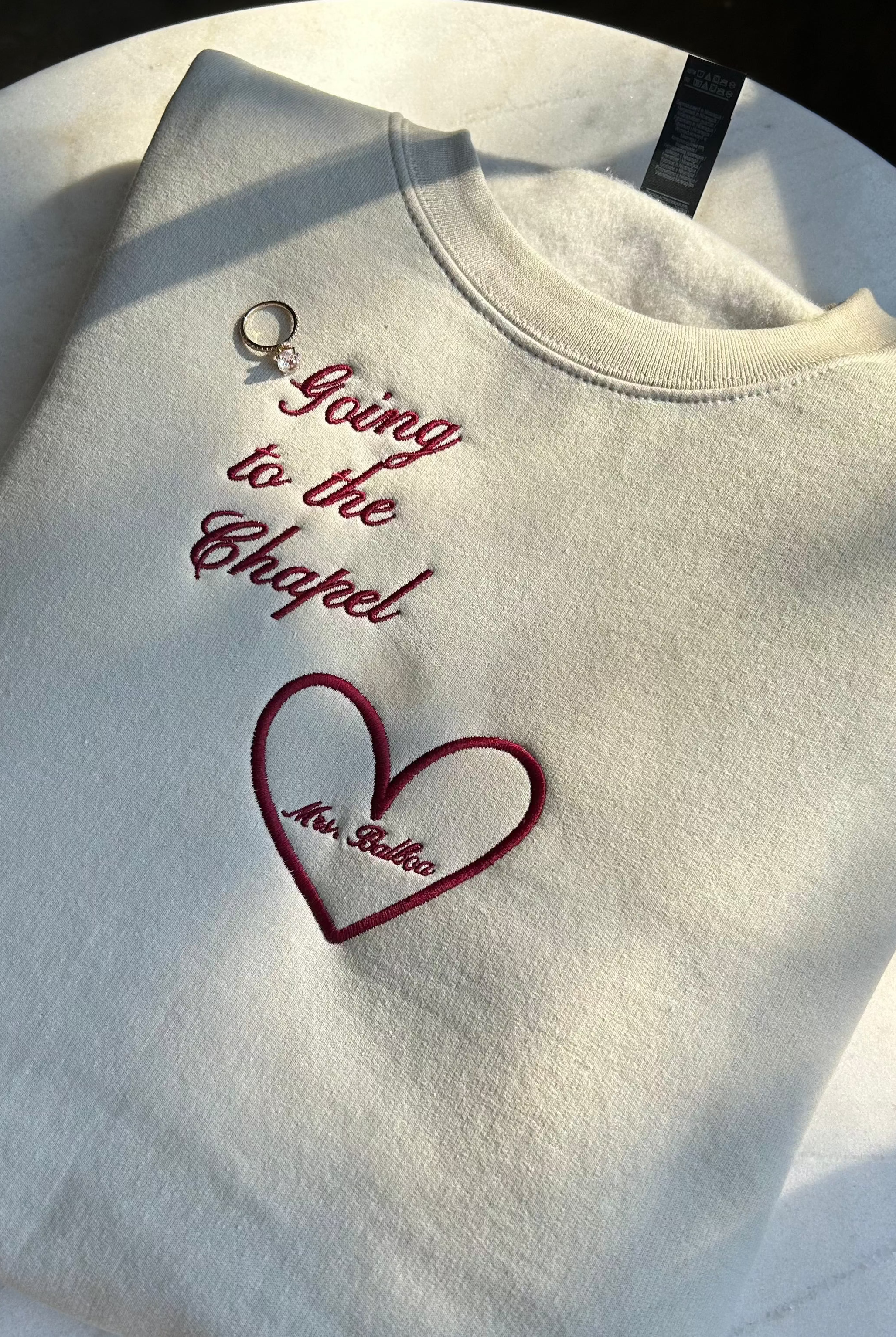 A beige embroidered crewneck sweatshirt with the phrase 'Going to the Chapel' and a heart design, using wine-colored thread.