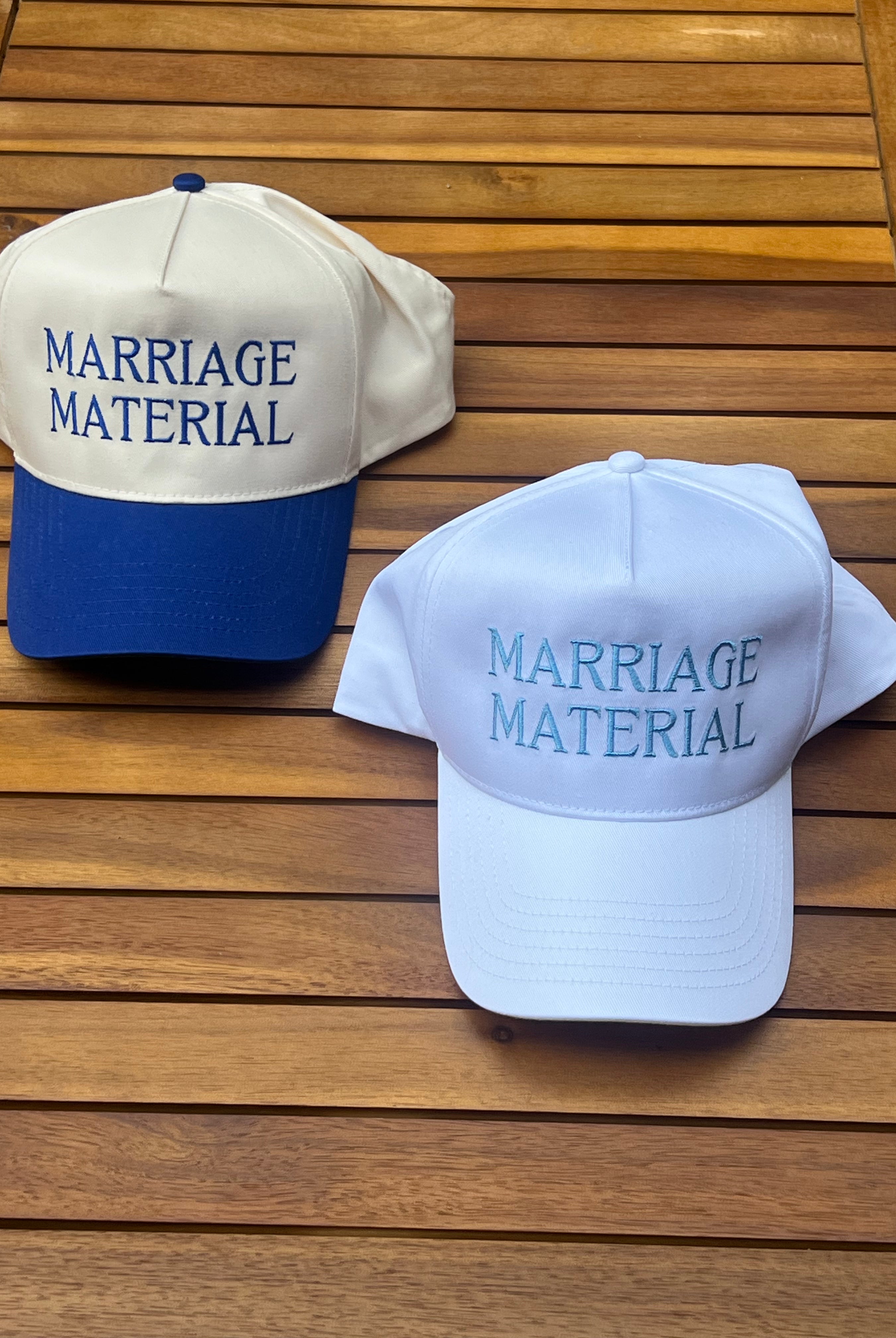 Two baseball caps with 'MARRIAGE MATERIAL' text on a wooden surface