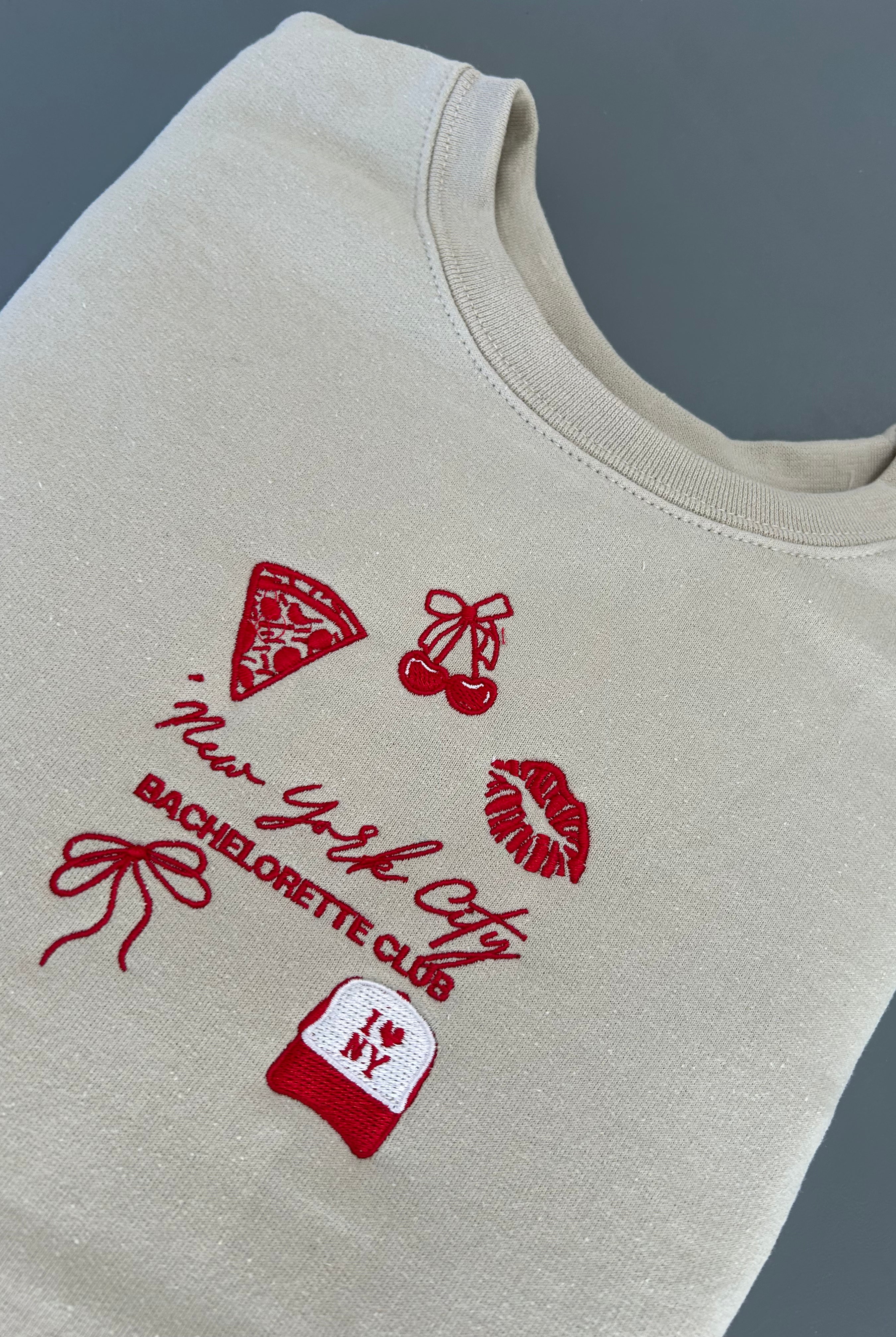 A beige embroidered sweatshirt with NYC-themed designs including a pizza slice, high heels, and a bow, along with the text 'New York City Bachelorette Club'.