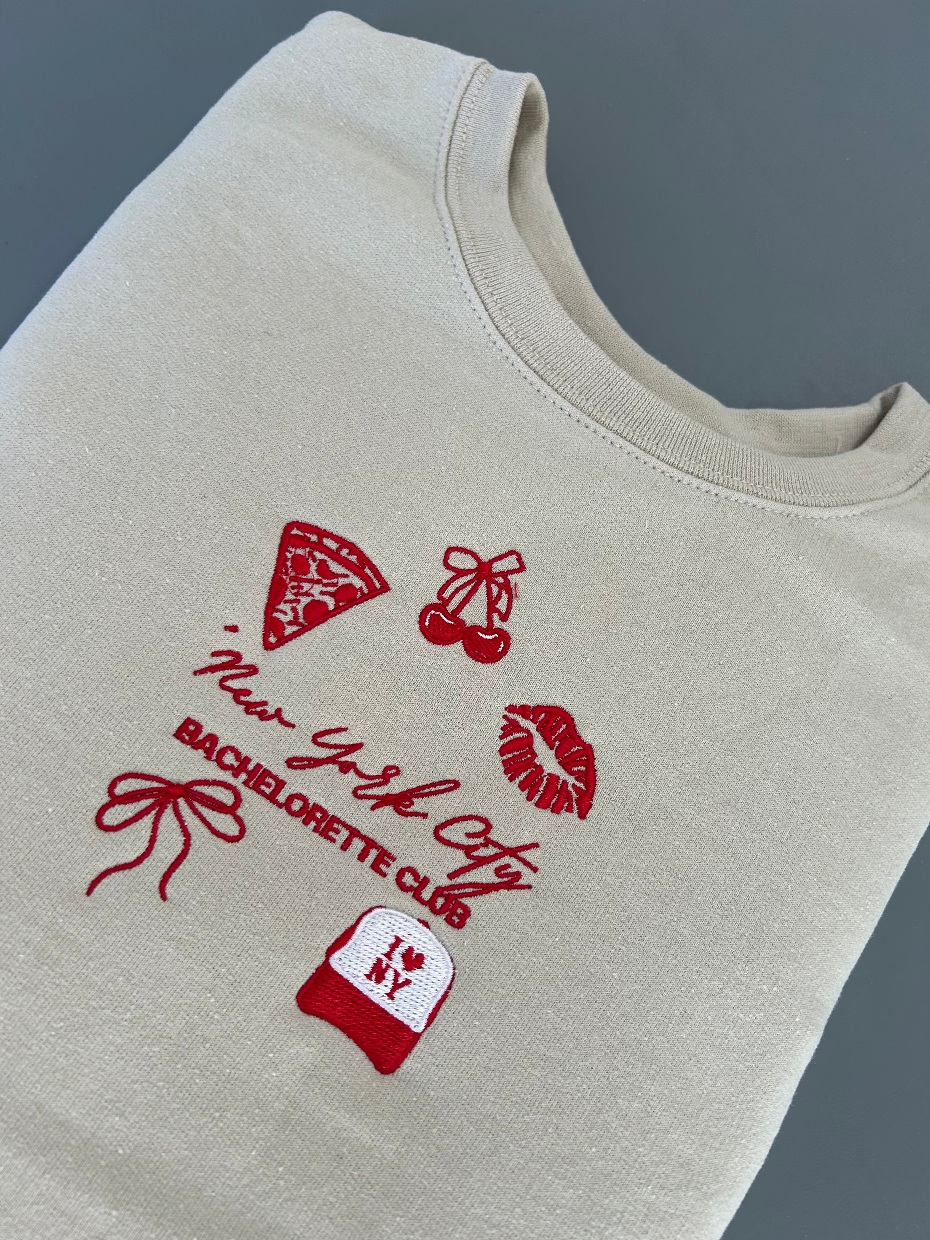 A beige embroidered sweatshirt with NYC-themed designs including a pizza slice, high heels, and a bow, along with the text 'New York City Bachelorette Club'.