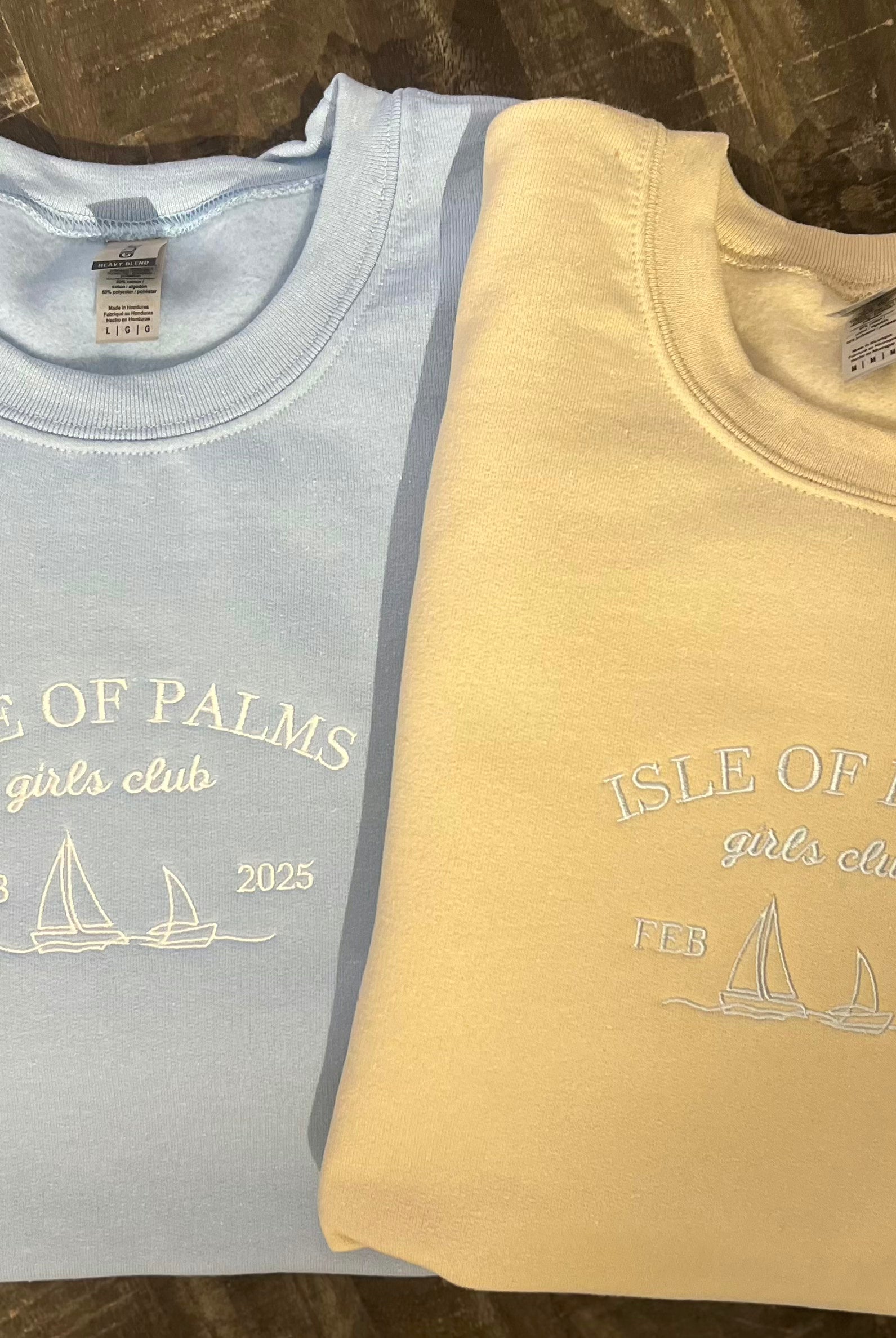 Two crewneck sweatshirts, one in blue and one in a beige color, each with a embroidered sailboat design and the text 'ISLE OF PALMS YACHT CLUB'.