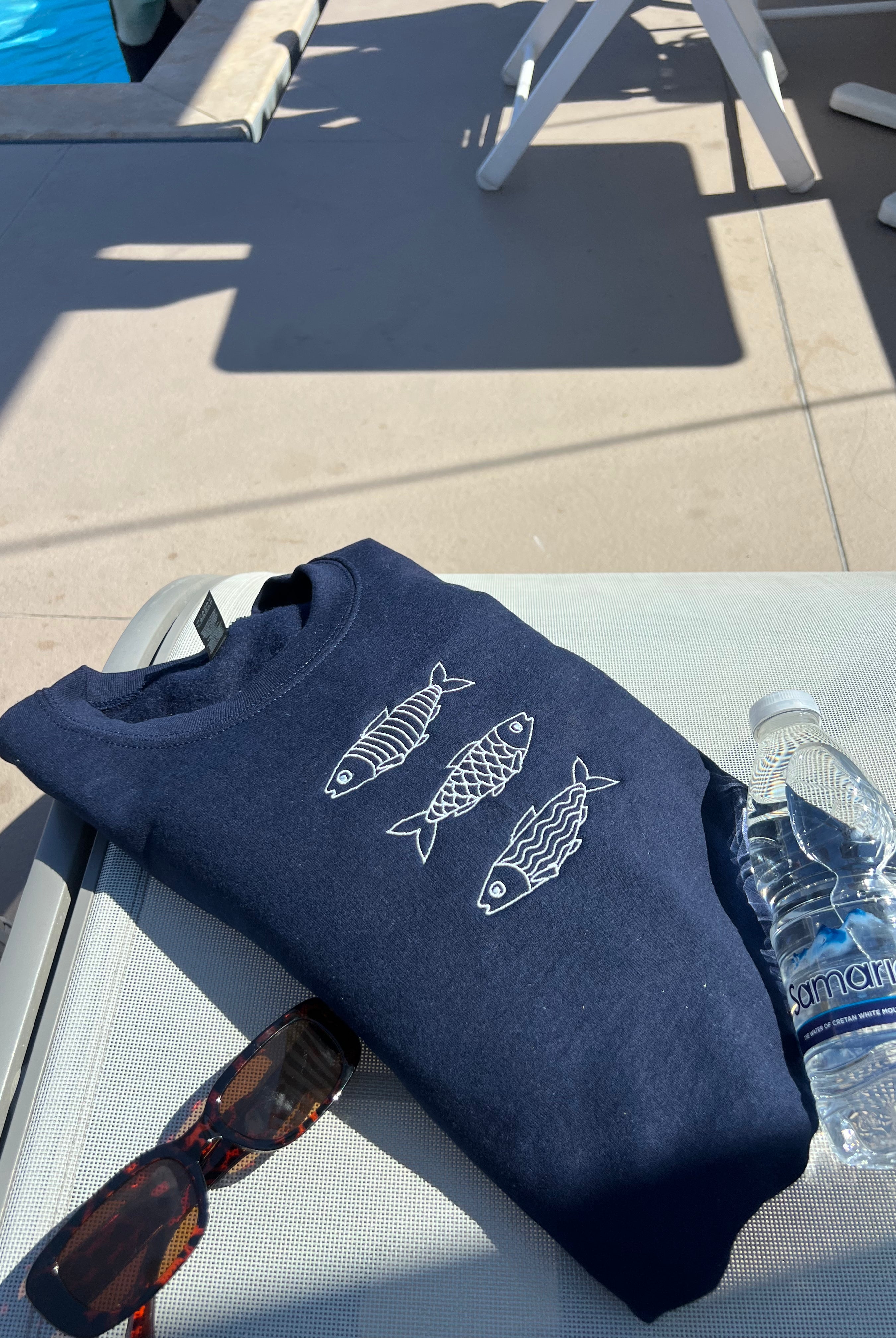 Navy blue sweatshirt with fish designs, sunglasses, and a bottle of water on a chair by a poolside.