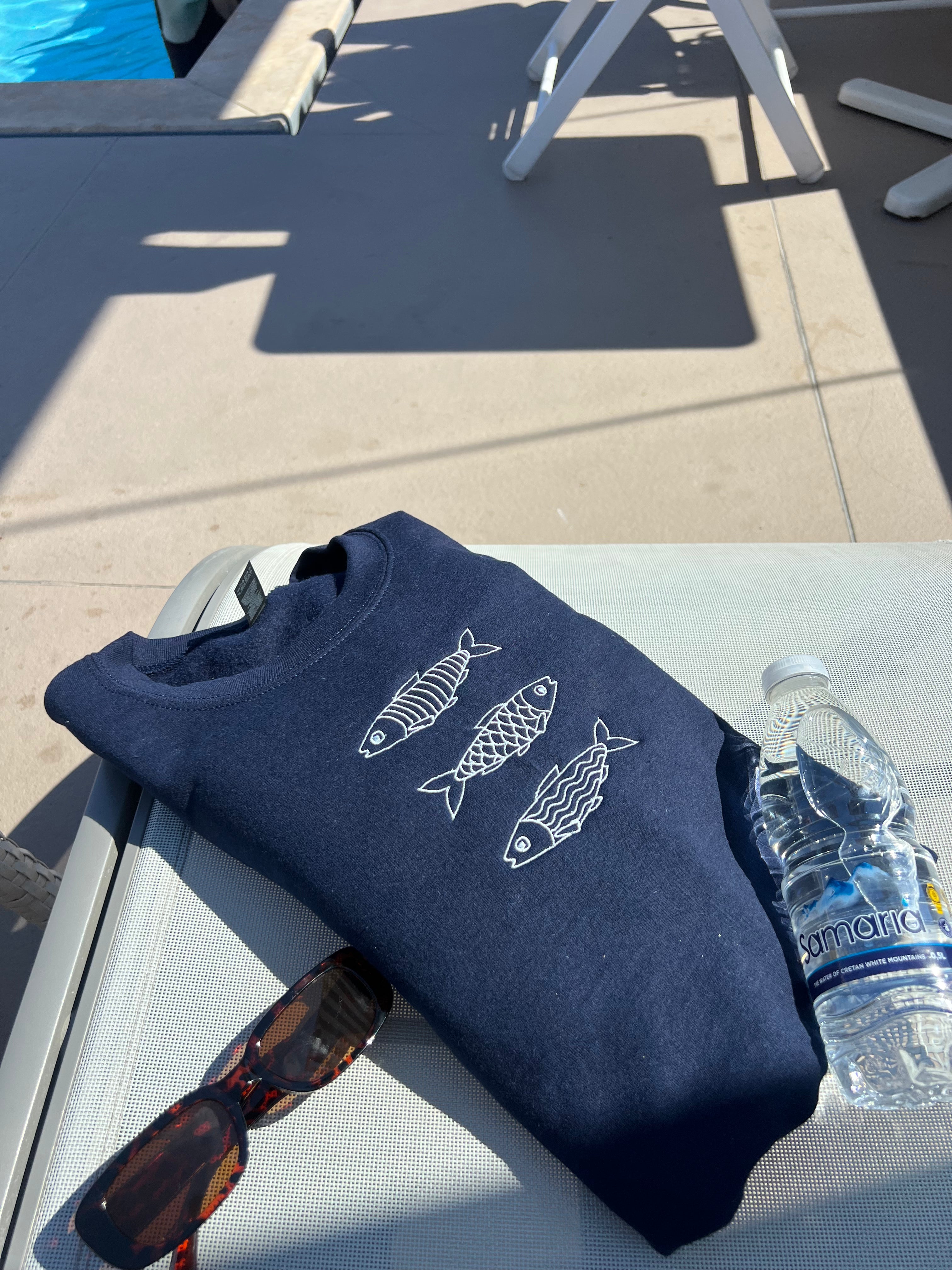 Navy blue sweatshirt with fish designs, sunglasses, and a bottle of water on a chair by a poolside.