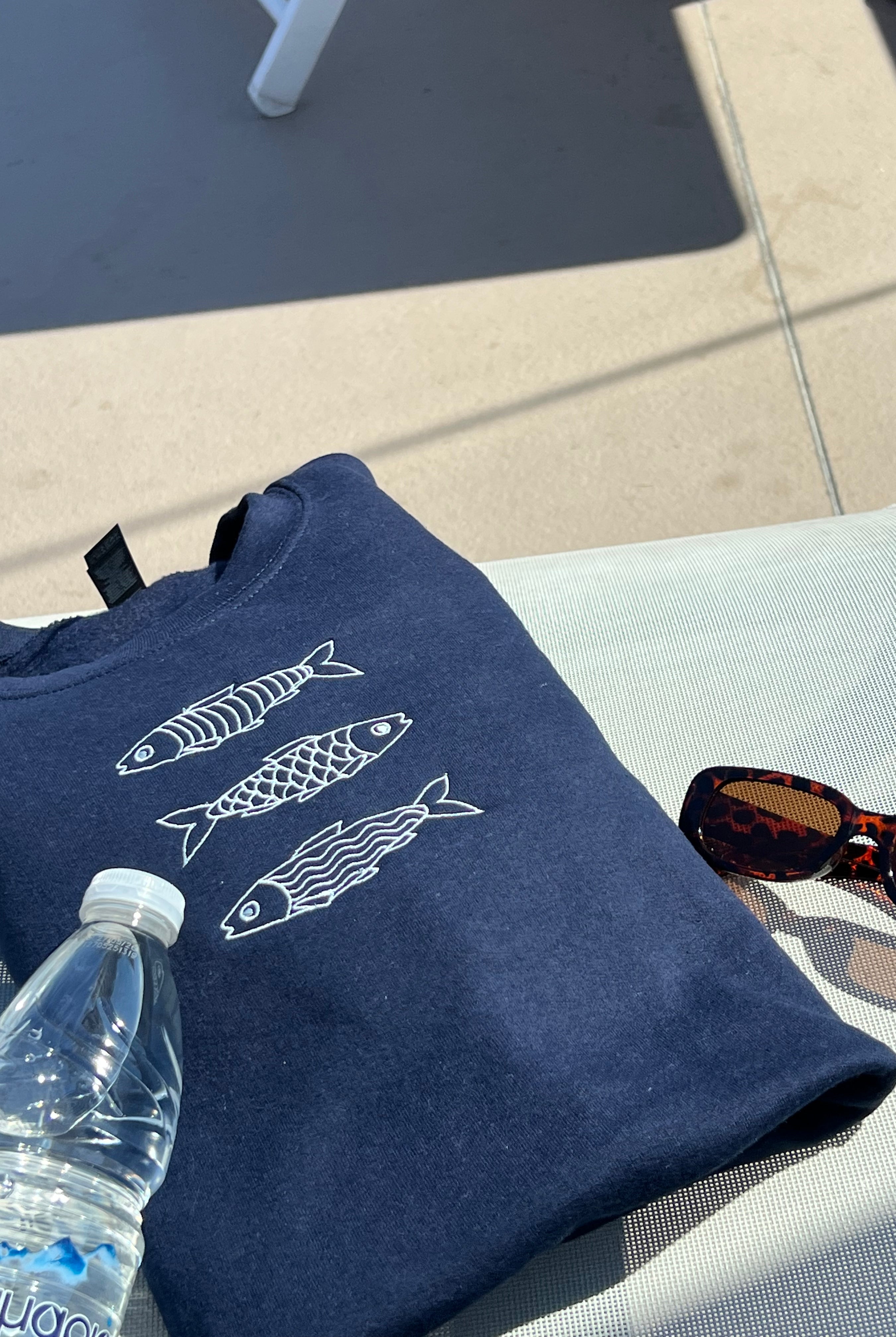 Navy blue sweatshirt with fish designs on a white surface next to a water bottle and sunglasses.