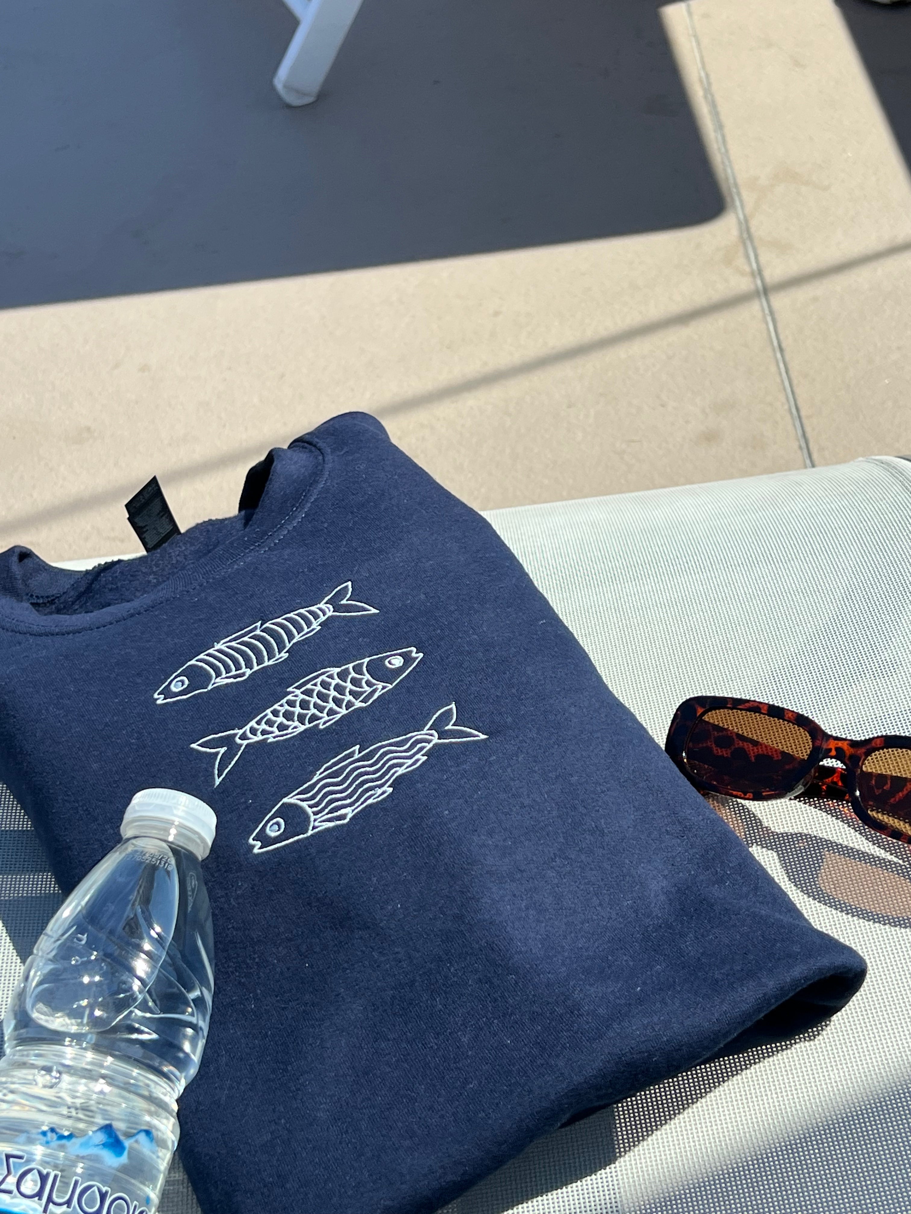 Navy blue sweatshirt with fish designs on a white surface next to a water bottle and sunglasses.