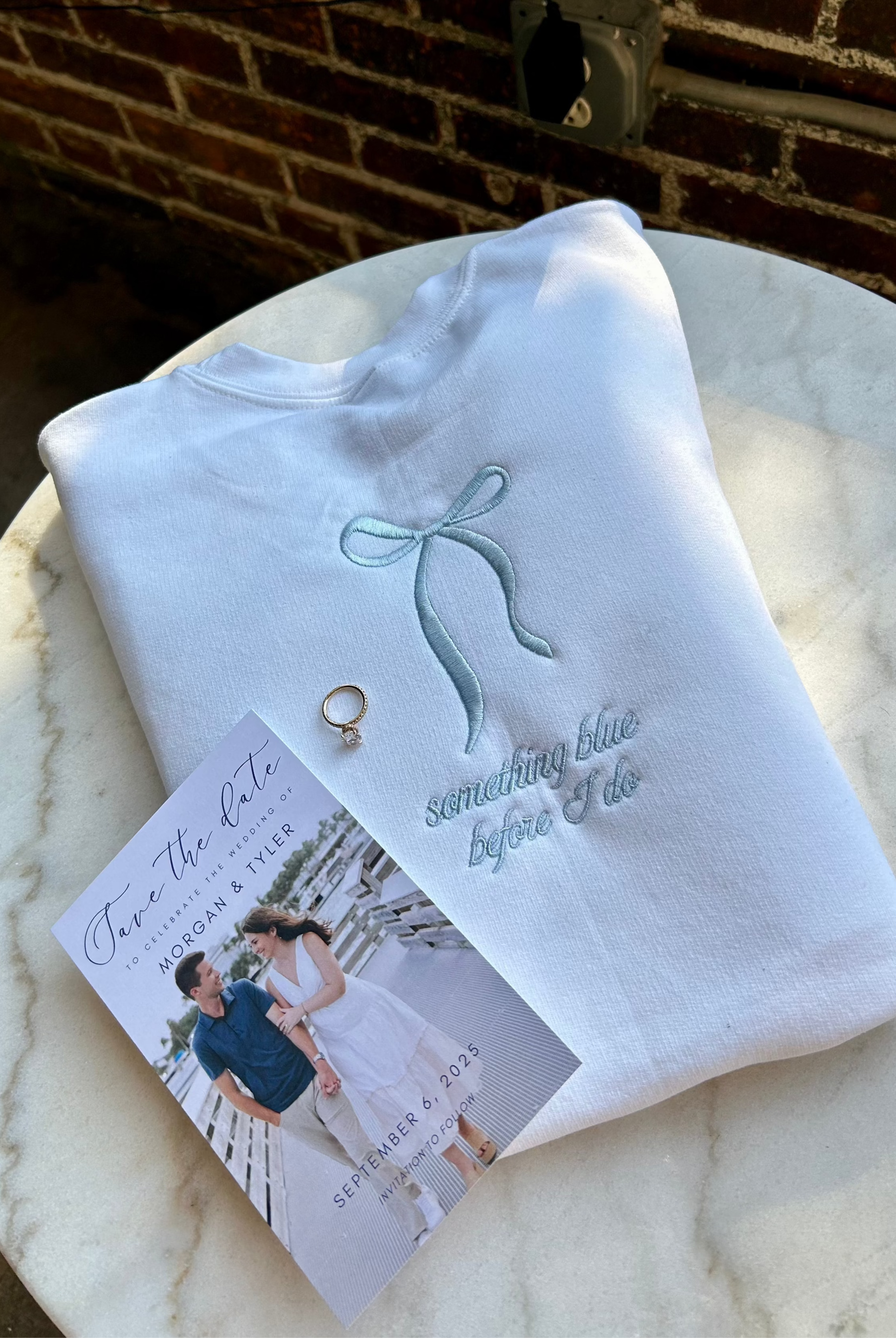 A white crewneck sweatshirt with light blue thread embroidery in the shape of a bow and text that reads 'something blue before i do'. There is also a small heart shape next to the text.