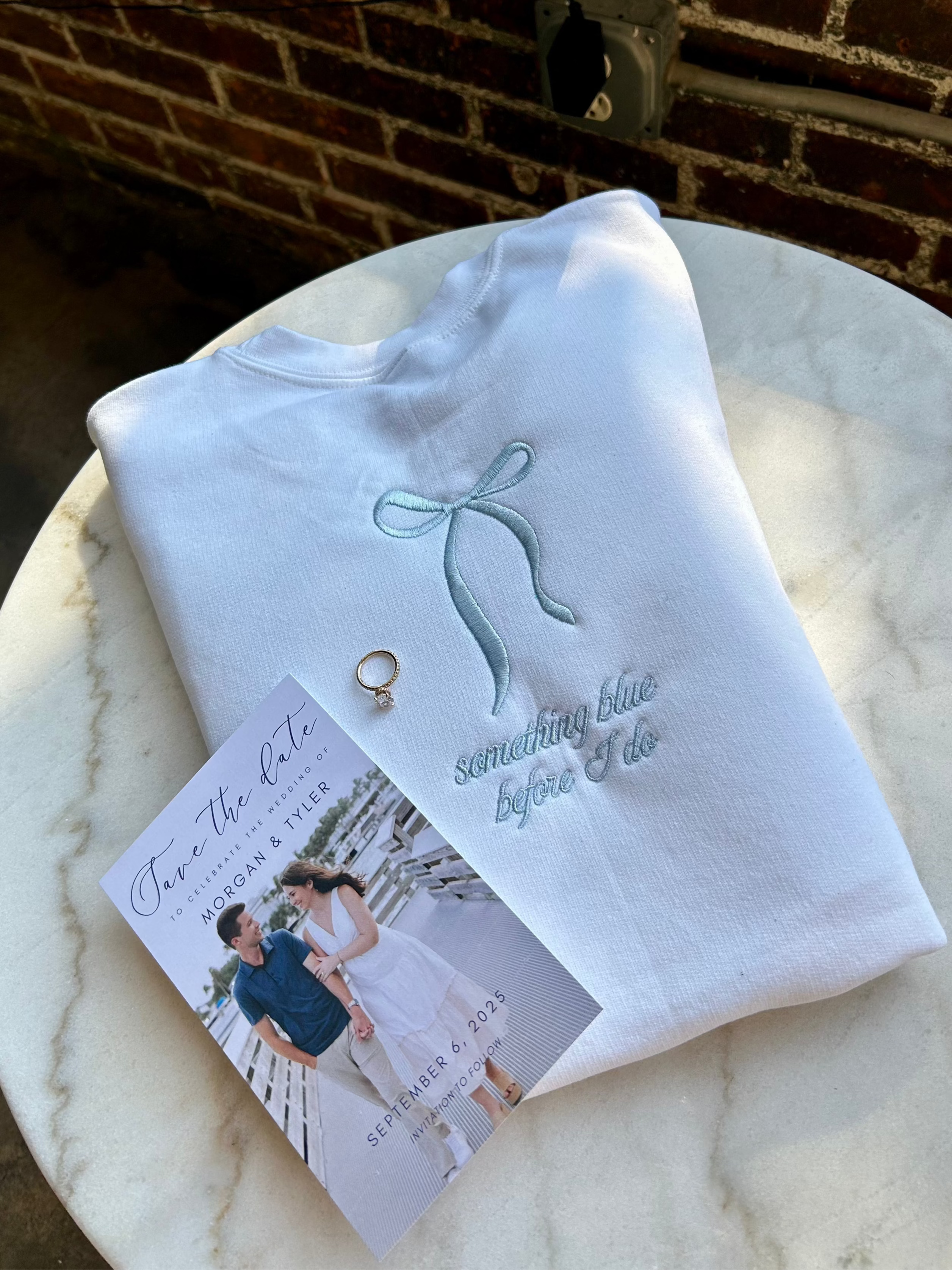 A white crewneck sweatshirt with light blue thread embroidery in the shape of a bow and text that reads 'something blue before i do'. There is also a small heart shape next to the text.