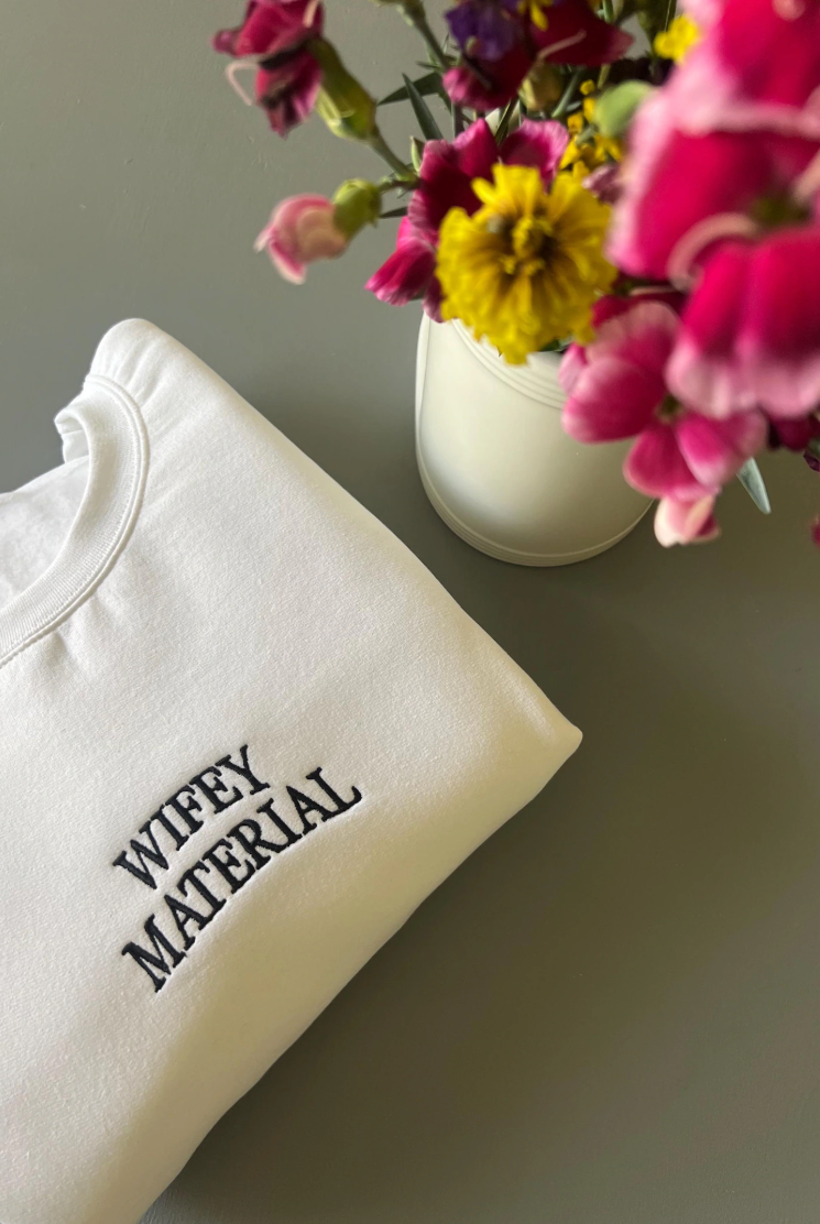 A white embroidered crewneck sweatshirt with the phrase 'Wifey Material' written in black thread, displayed next to a vase of flowers.