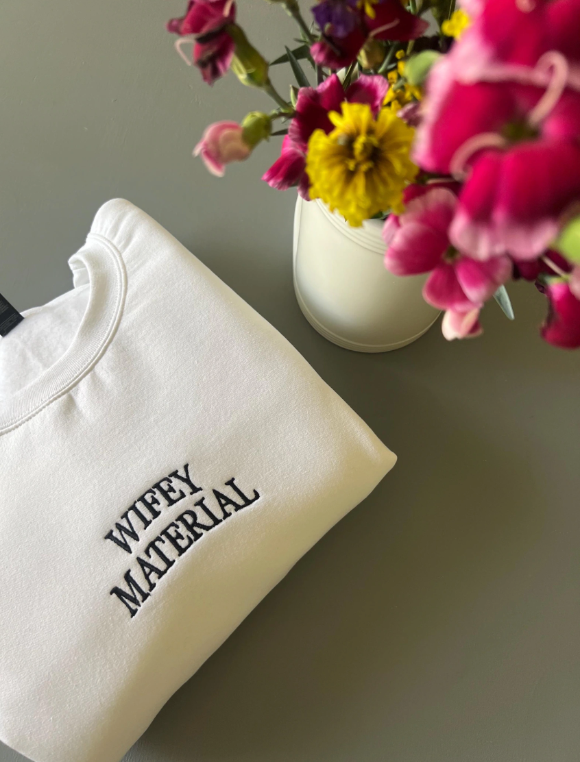 A white embroidered crewneck sweatshirt with the phrase 'Wifey Material' written in black thread, displayed next to a vase of flowers.