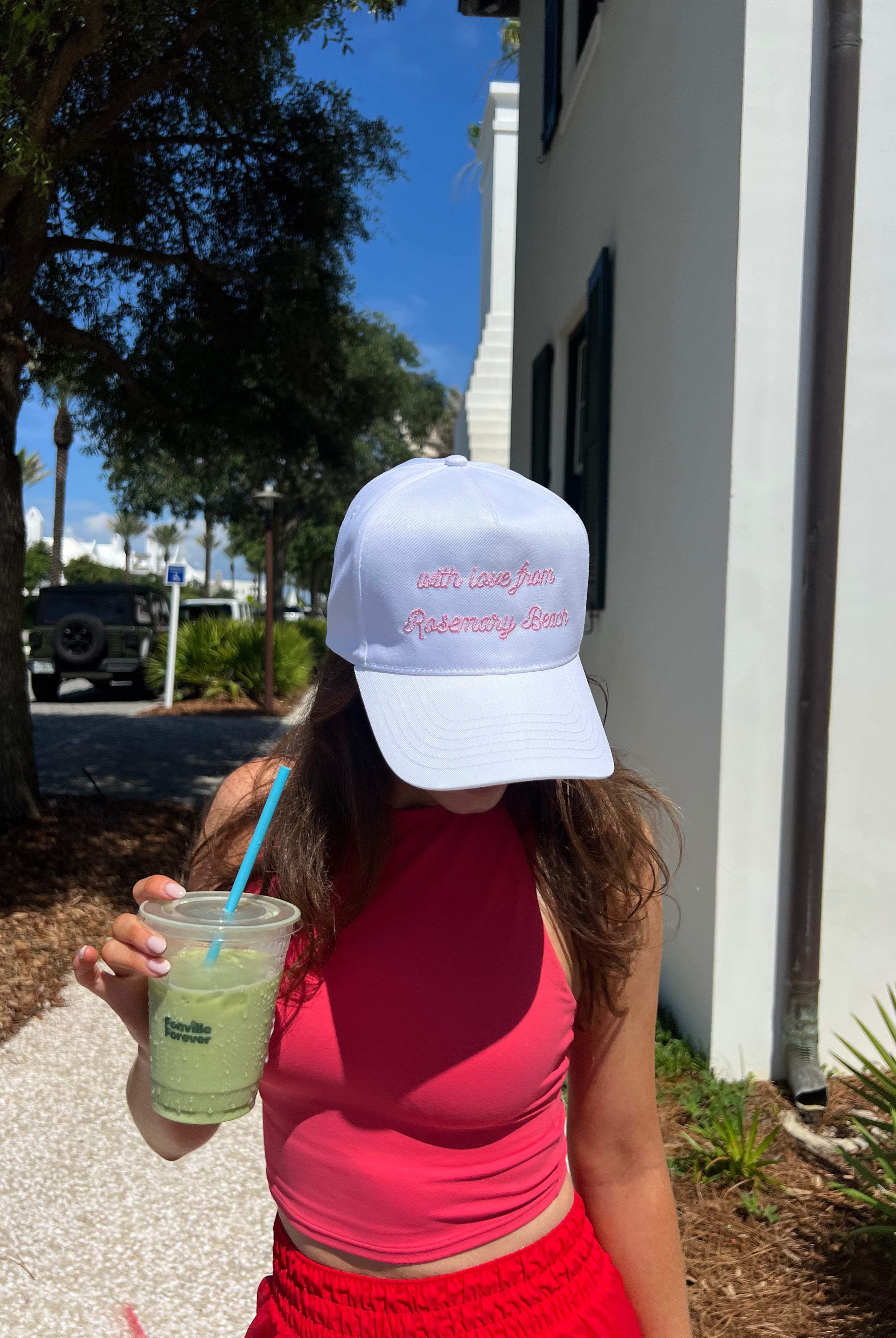 Person wearing a white cap with text, holding a drink, standing outdoors.
