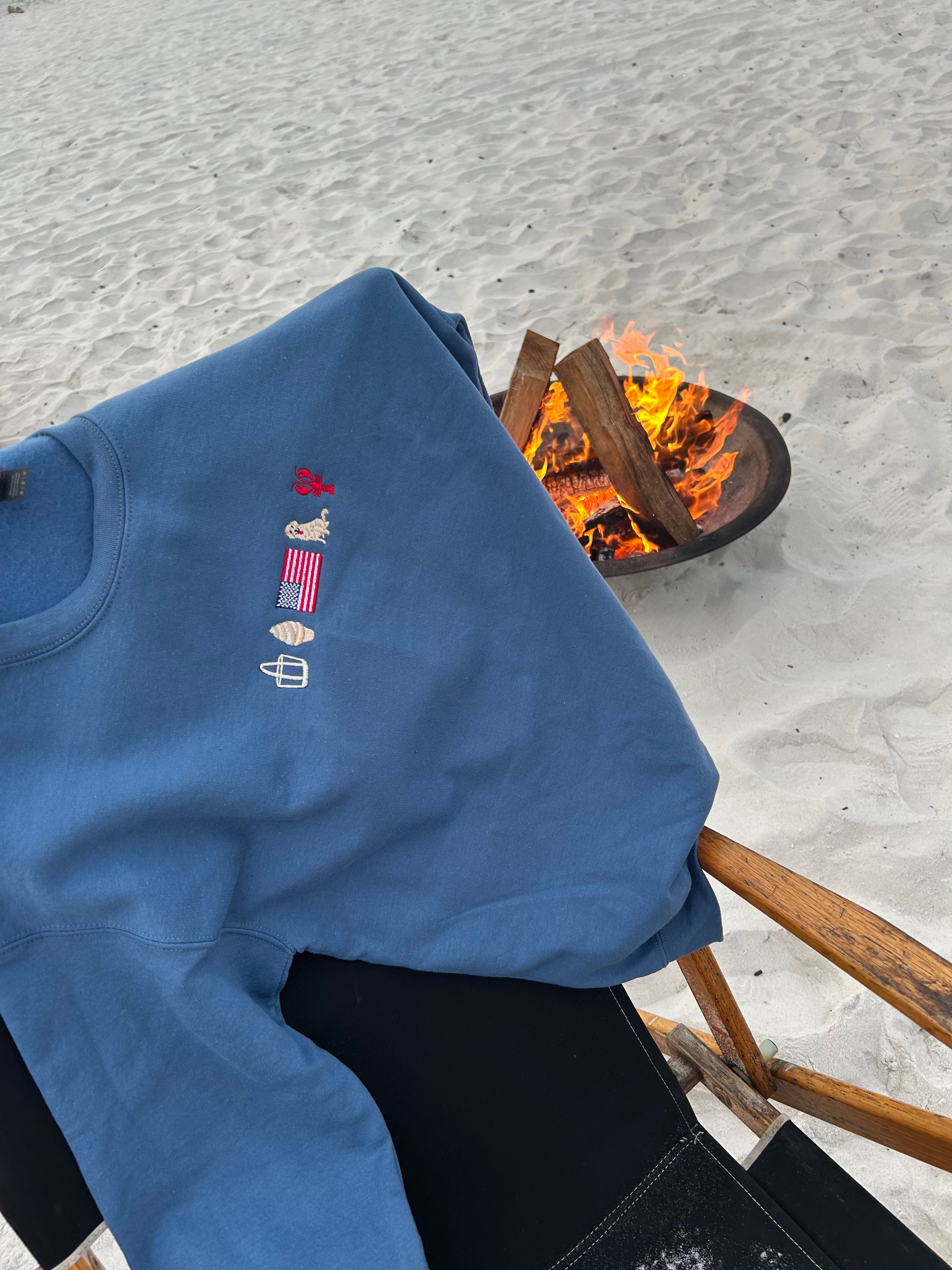 Blue sweatshirt with embroidered designs on a beach chair next to a campfire.