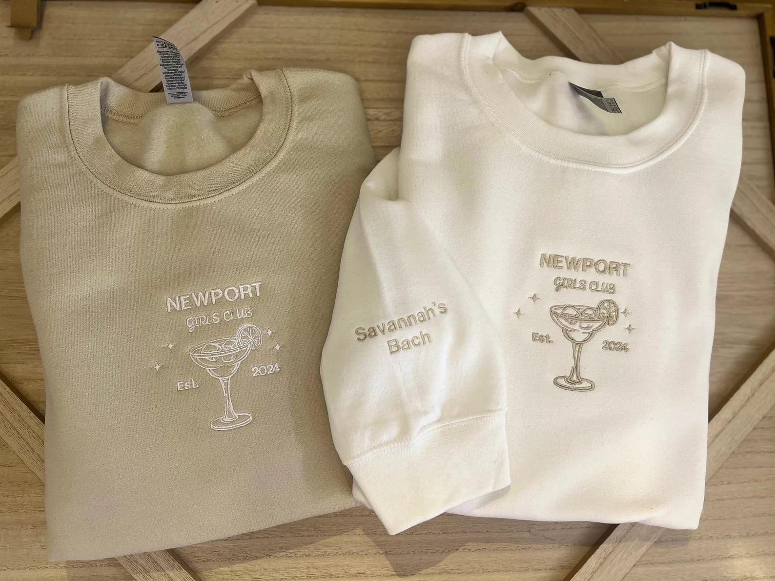 Two folded embroidered crewneck sweatshirts, one beige and one white, with a custom margarita glass design and text on them.