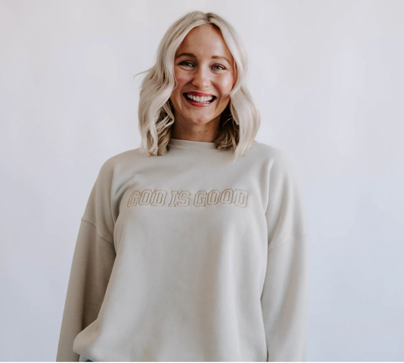 A person wearing a dust-colored sweatshirt with 'GOOD IS GOOD' embroidered on the front.