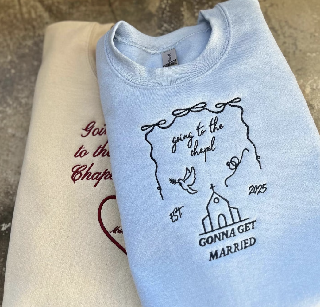Light blue embroidered sweatshirt with text and design elements indicating a bridal theme, displayed next to a beige item, possibly another sweatshirt or a hat.