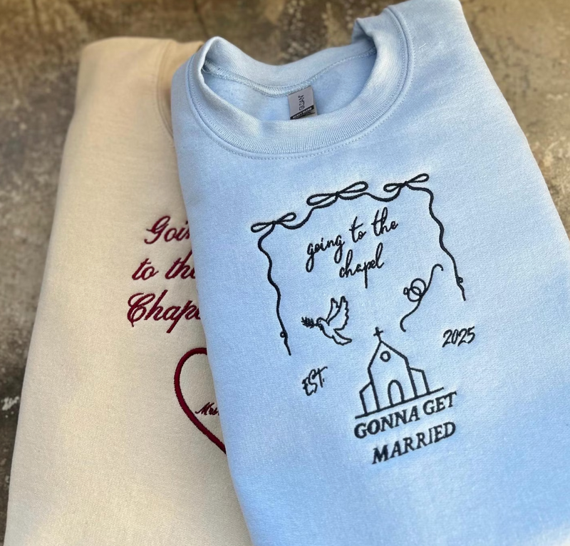 Light blue embroidered sweatshirt with text and design elements indicating a bridal theme, displayed next to a beige item, possibly another sweatshirt or a hat.