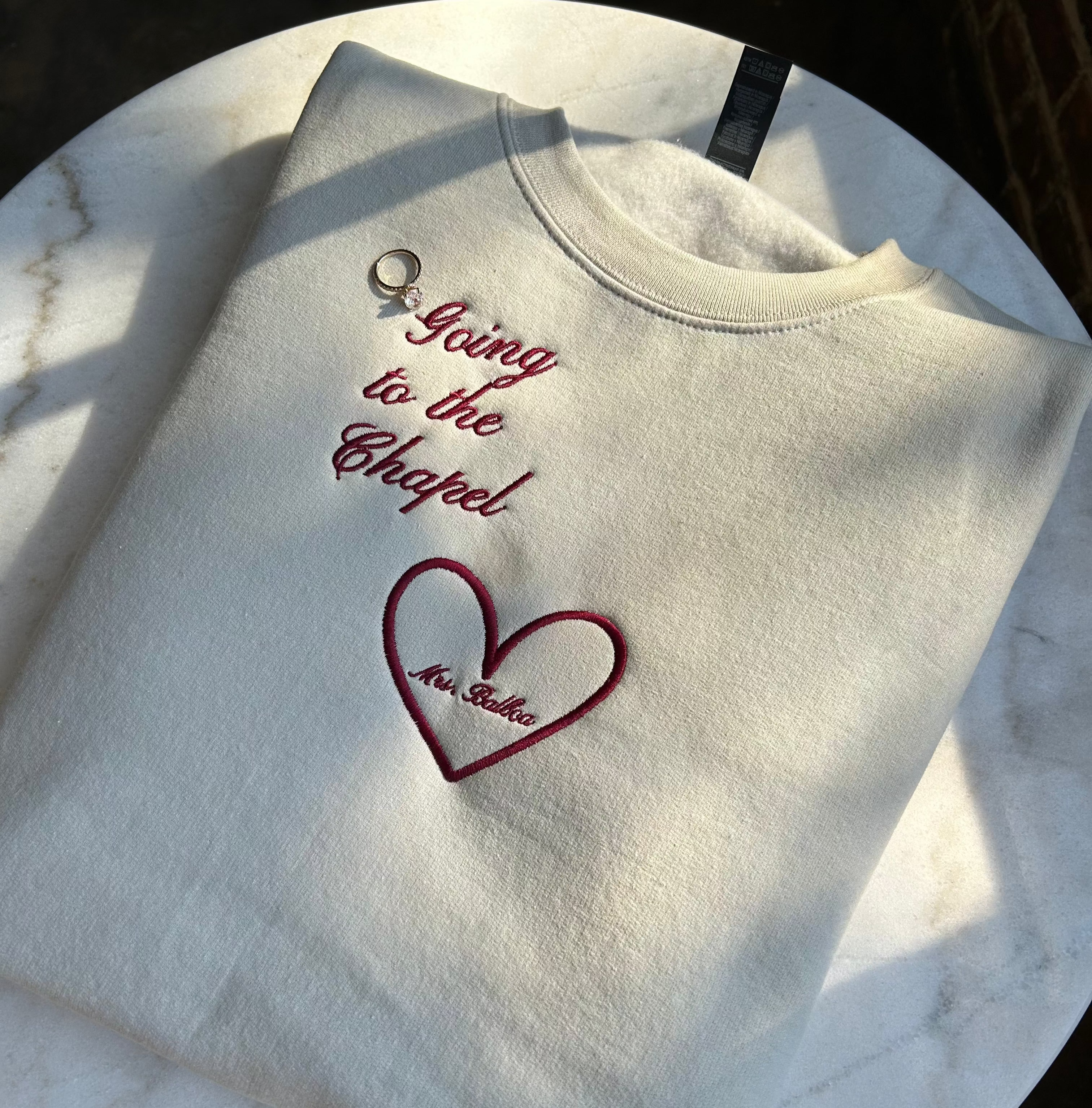 A beige embroidered crewneck sweatshirt with the phrase 'Going to the Chapel' and a heart design, using wine-colored thread.