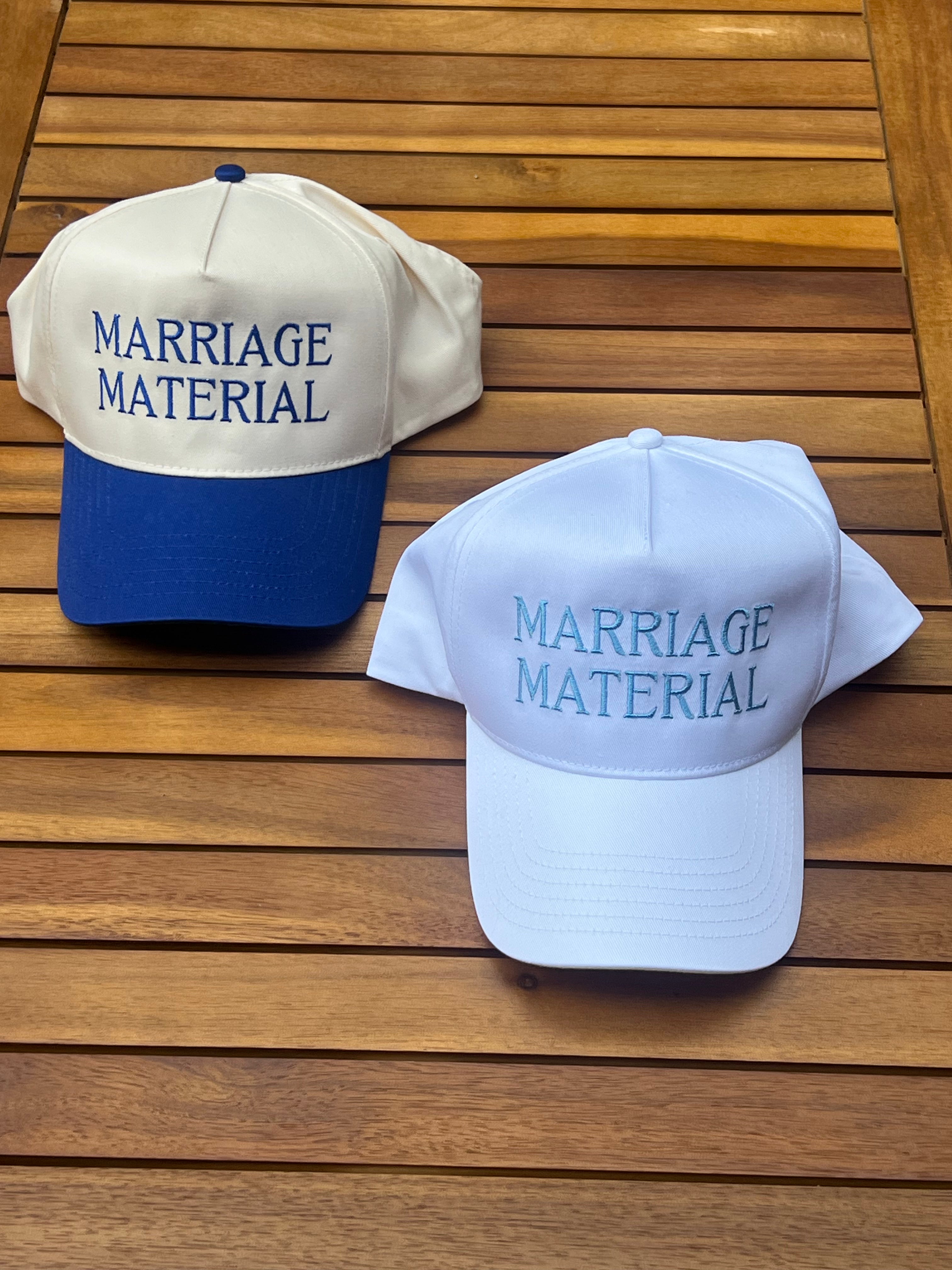 Two baseball caps with 'MARRIAGE MATERIAL' text on a wooden surface
