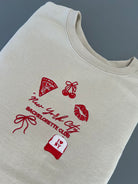 A beige embroidered sweatshirt with NYC-themed designs including a pizza slice, high heels, and a bow, along with the text 'New York City Bachelorette Club'.