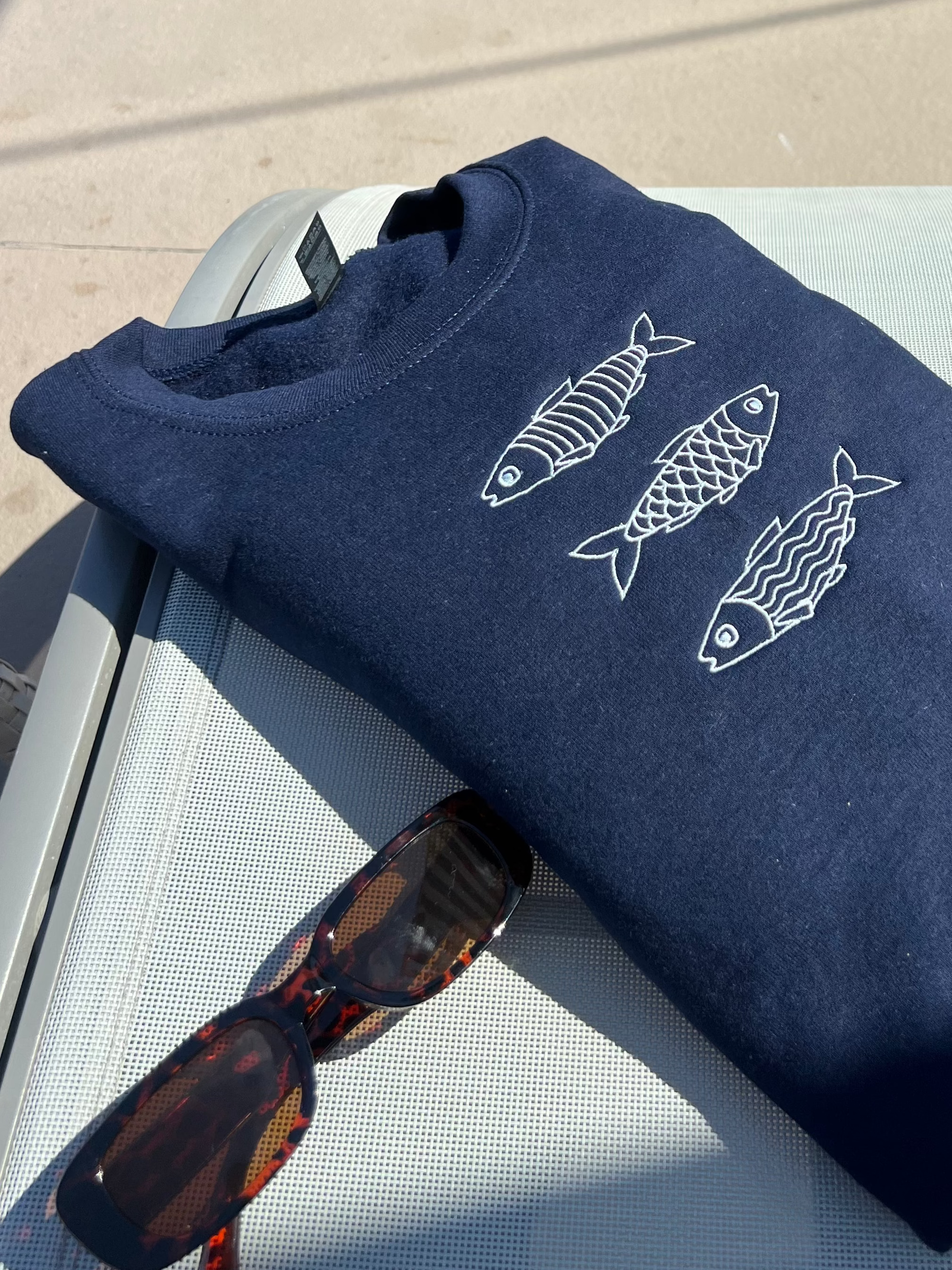 Navy blue sweatshirt with fish designs, sunglasses, and a bottle of water on a chair by a poolside.