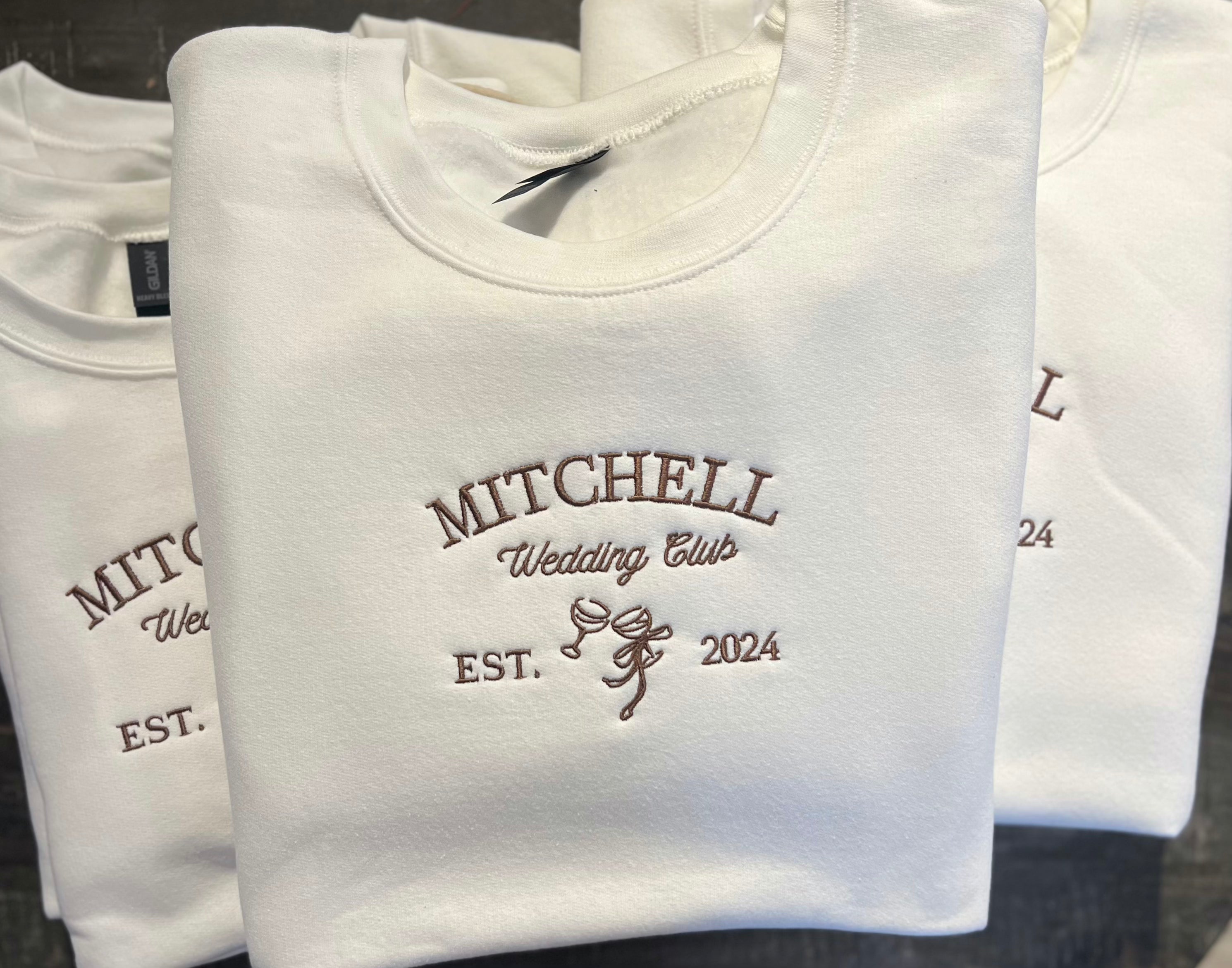 A collection of white embroidered sweatshirts with the text 'MITCHELL Wedding Club EST. 2024' on them.