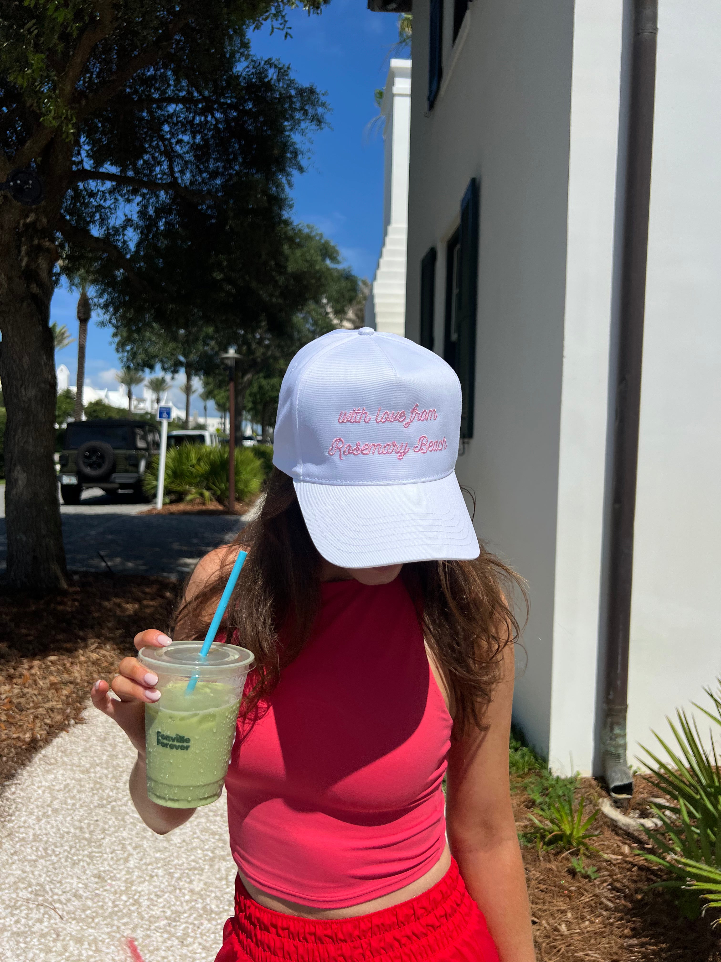 Person wearing a white cap with text, holding a drink, standing outdoors.